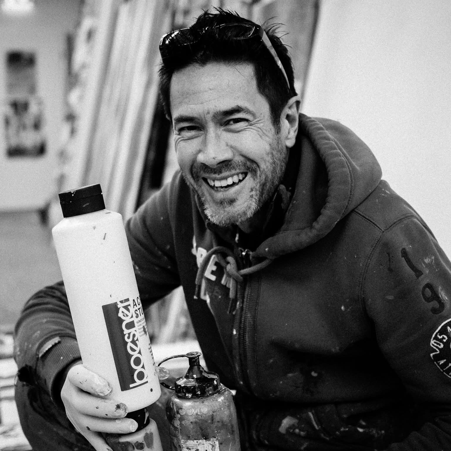 Frank Jons à l'atelier, souriant, tenant un flacon de peinture acrylique
